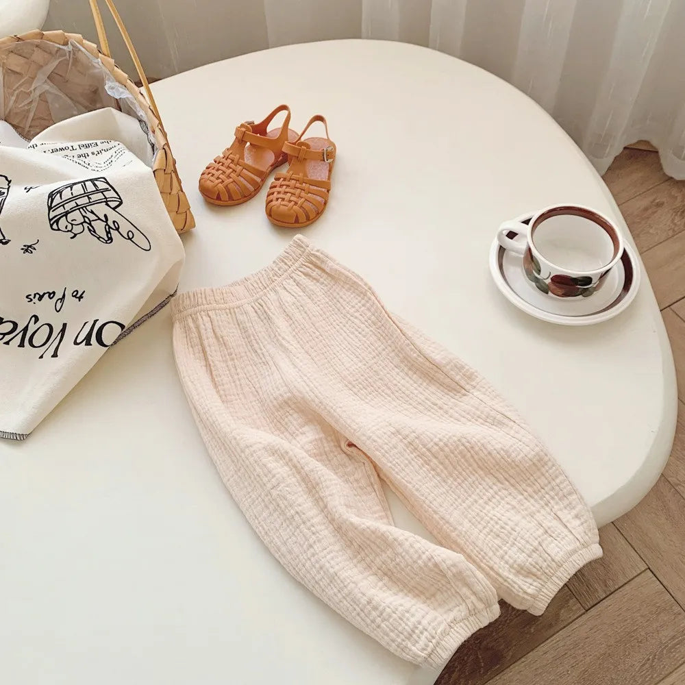 Light pink baby pants on a white surface with a basket, shoes, and cup in the background.