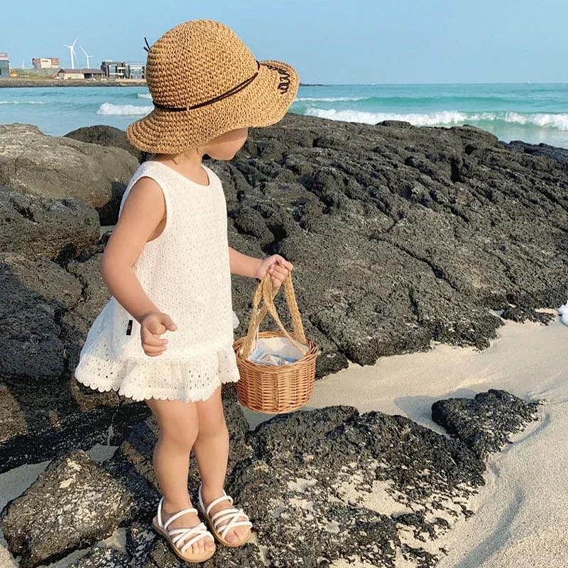 Child in a white dress and straw hat standing on rocky beach with ocean in background